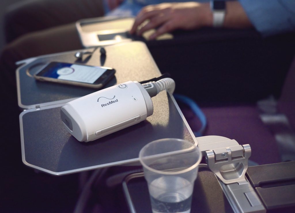 Resmed AirMini AutoSet Portable CPAP device placed on airplane tray table next to a smartphone and water cup
