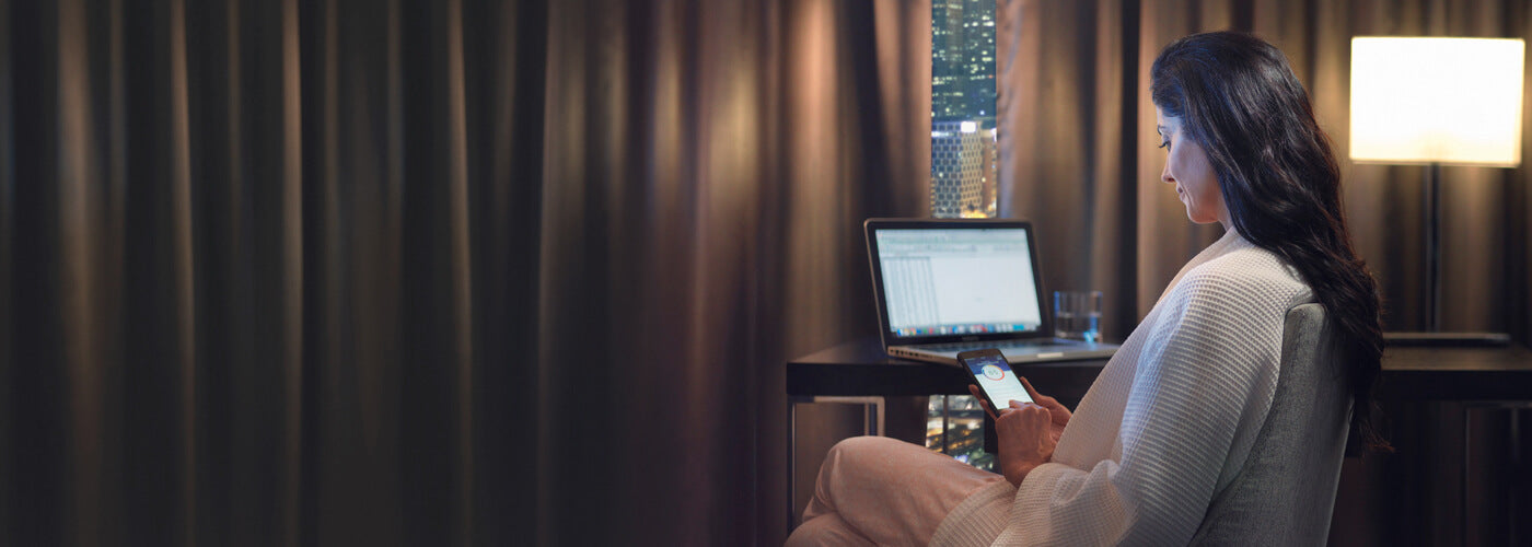 Woman using laptop and phone in hotel room with Resmed AirMini AutoSet Portable CPAP beside her
